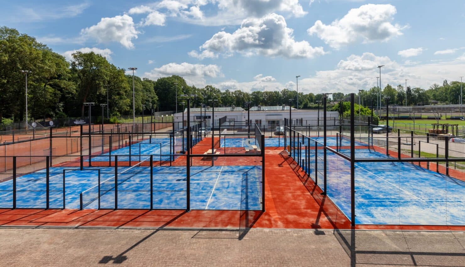 Outdoor padelcomplex met meerdere courts omgeven door zwart gaas en glas, blauwe en rode ondergronden, clubhuis zichtbaar.