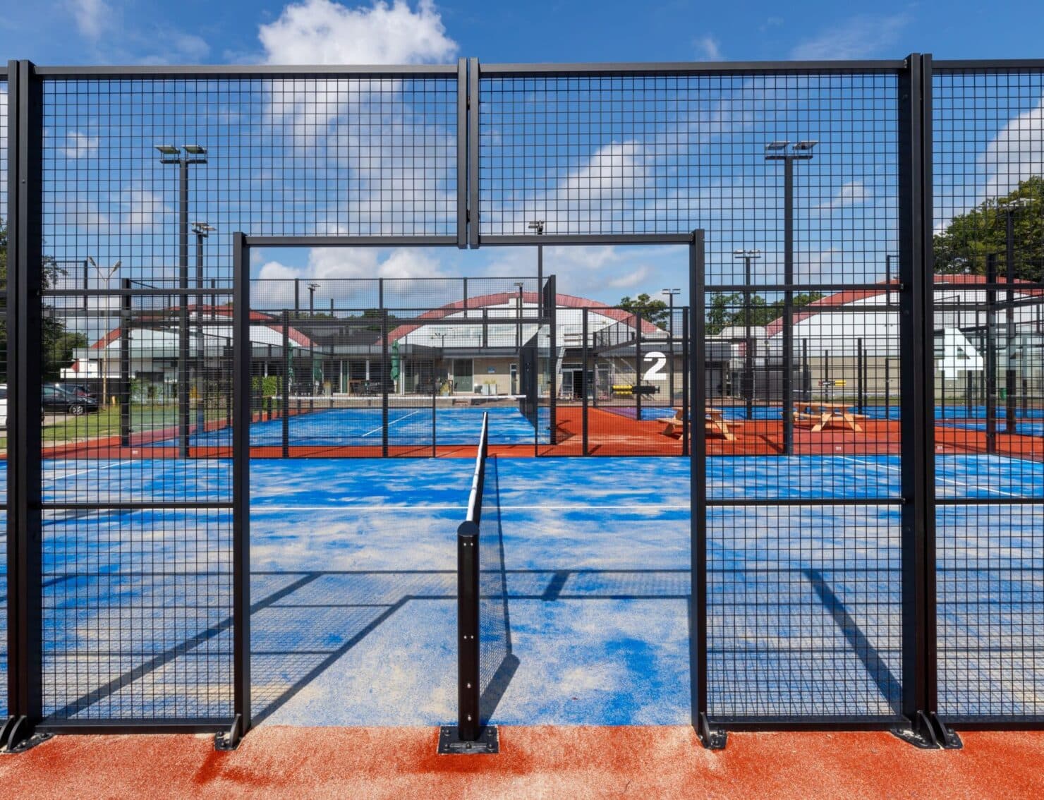 Outdoor padelcomplex met blauwe kunstgrasbanen achter zwart gaaswerk, clubhuis en verlichting zichtbaar.