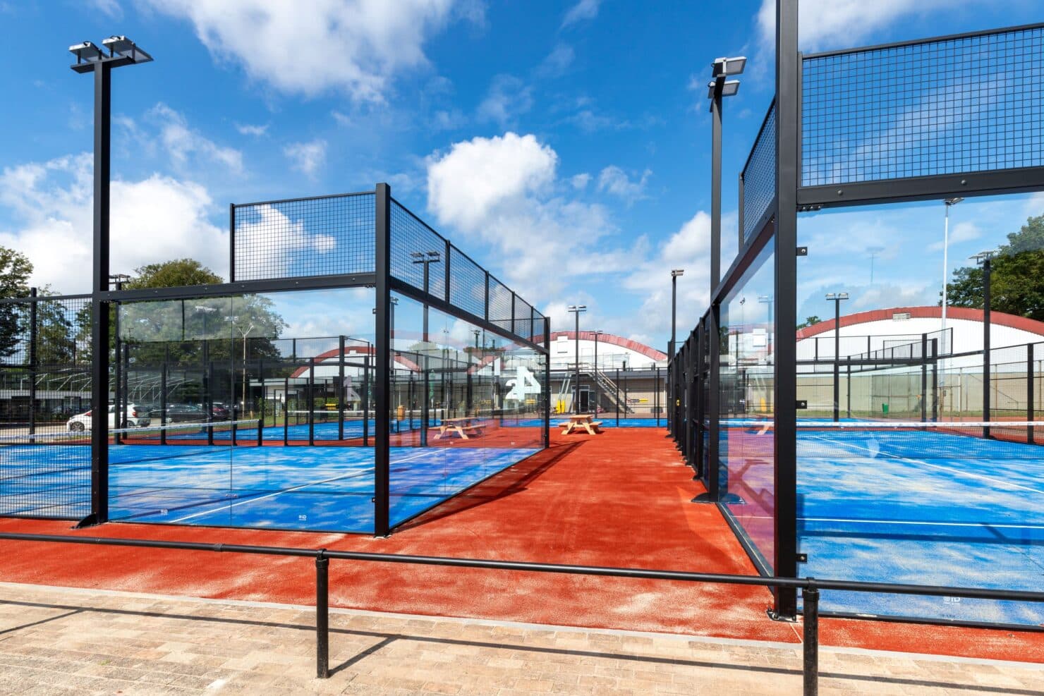 Outdoor padelcomplex met meerdere blauwe velden omgeven door zwart gaas, rode ondergrond, daglicht.