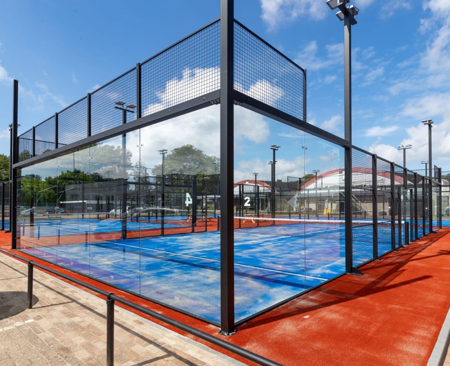 Moderne outdoor padeltennisbaan met blauwe veldoppervlak, zwart metalen kooi-constructie en verlichting.
