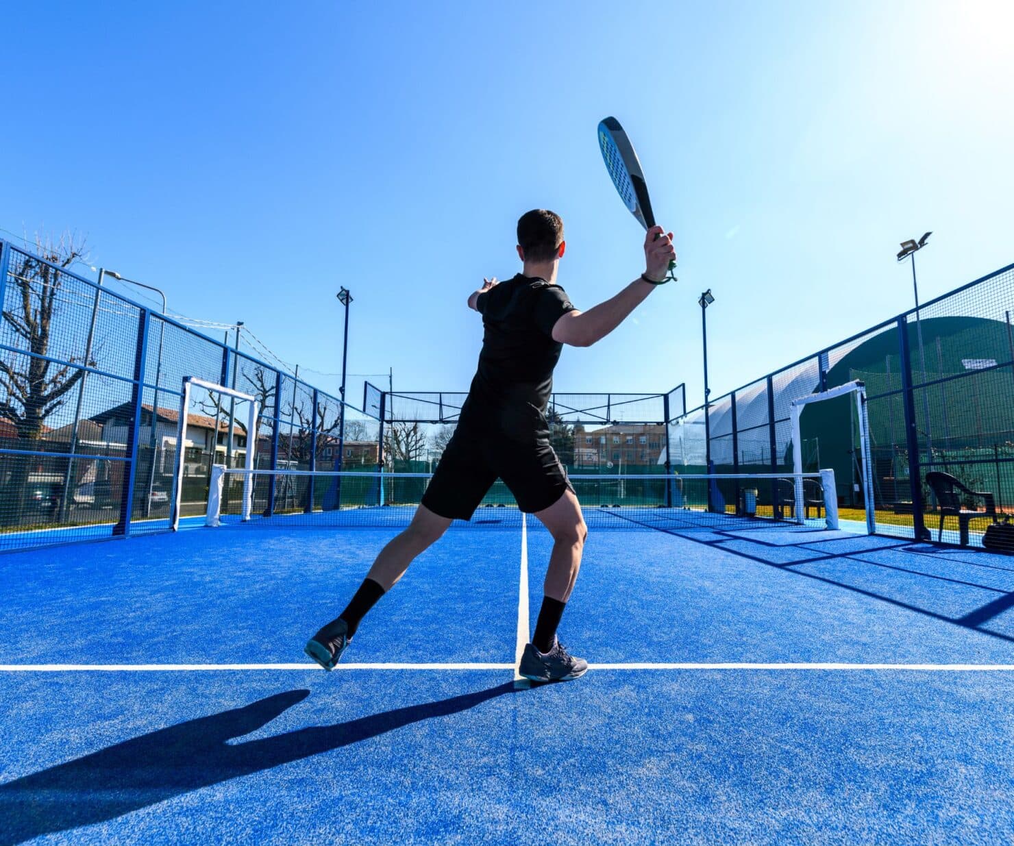 Padelspeler in actiepositie op blauwe buitenbaan met zwart meshkooi omheing.