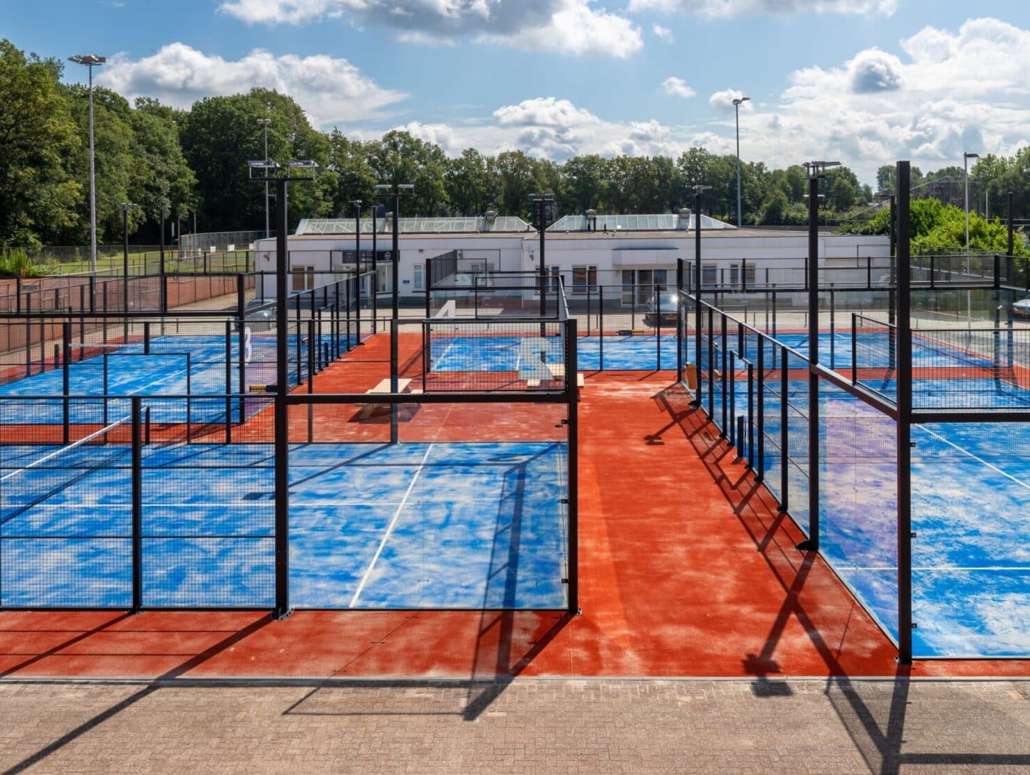 Moderne outdoor padelcomplex met meerdere blauwe en rode courts omgeven door zwart gaas en glazen wanden.
