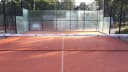 Outdoor padel court met rode ondergrond, omsloten door glazen en metalen mesh wanden.