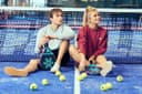 Twee padelspelers zitten op blauw hardcourt met tennisballen en rackets bij het net