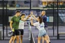 Vier padelspelers geven high-fives op een indoorbaan met glazen achterwand en netwerk omheining.