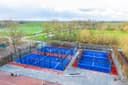 Luchtfoto van een moderne padelbaan met vier blauwe courts en rood zijvelden in een groen landschap.