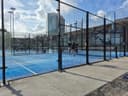 Outdoor padelbanencomplex met blauwe hardcourt velden, zwarte afscheidingen en stedelijke skyline.