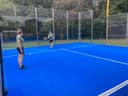 Twee spelers op een blauwe padelbaan met netting, outdoor club met bomen op achtergrond.