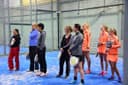 Groep vrouwen op blauwe padelbaan in overdekte sporthal met glazen wanden