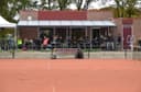 Padelbaan buiten met clubhuis, toeschouwers op terras, rode ondergrond zichtbaar.