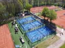 Luchtfoto van padelclub met drie blauwe banen, rode tennisbanen en groen terrein omgeven door bomen.