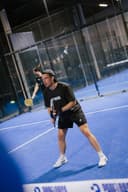 Padelspeler in zwart shirt op blauwe binnenbaan met glazen wanden en overhead verlichting.