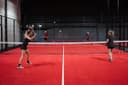 Twee speelsters spelen padel op een rood kunststofveld met netwerk omheining in overdekte baan.