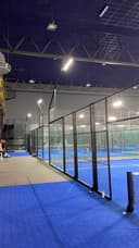 Binnenpadelbaancomplex met blauwe QuickPadel-ondergrond, zwart meshscherm en LED-verlichting.