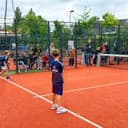 Outdoor padelcourt met oranje kunststofveld, omgeven door gaas en glaskooien, kinderen spelen match.